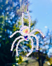 Close-up shot of a handmade Swarovski crystal spider suncatcher hanging outdoors. The faceted glass body and beaded iridescent legs shimmer brightly against a vibrant background of blue sky and sunny autumn trees.