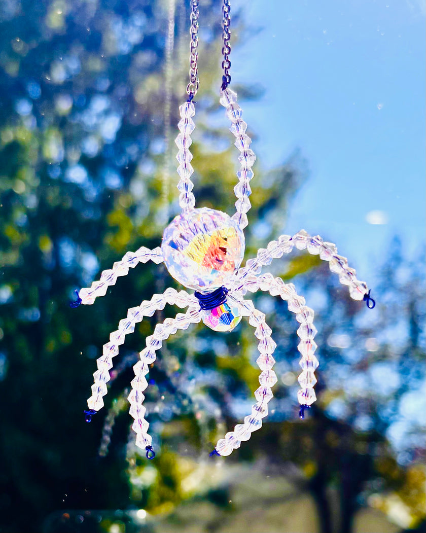 Close-up shot of a handmade Swarovski crystal spider suncatcher hanging outdoors. The faceted glass body and beaded iridescent legs shimmer brightly against a vibrant background of blue sky and sunny autumn trees.