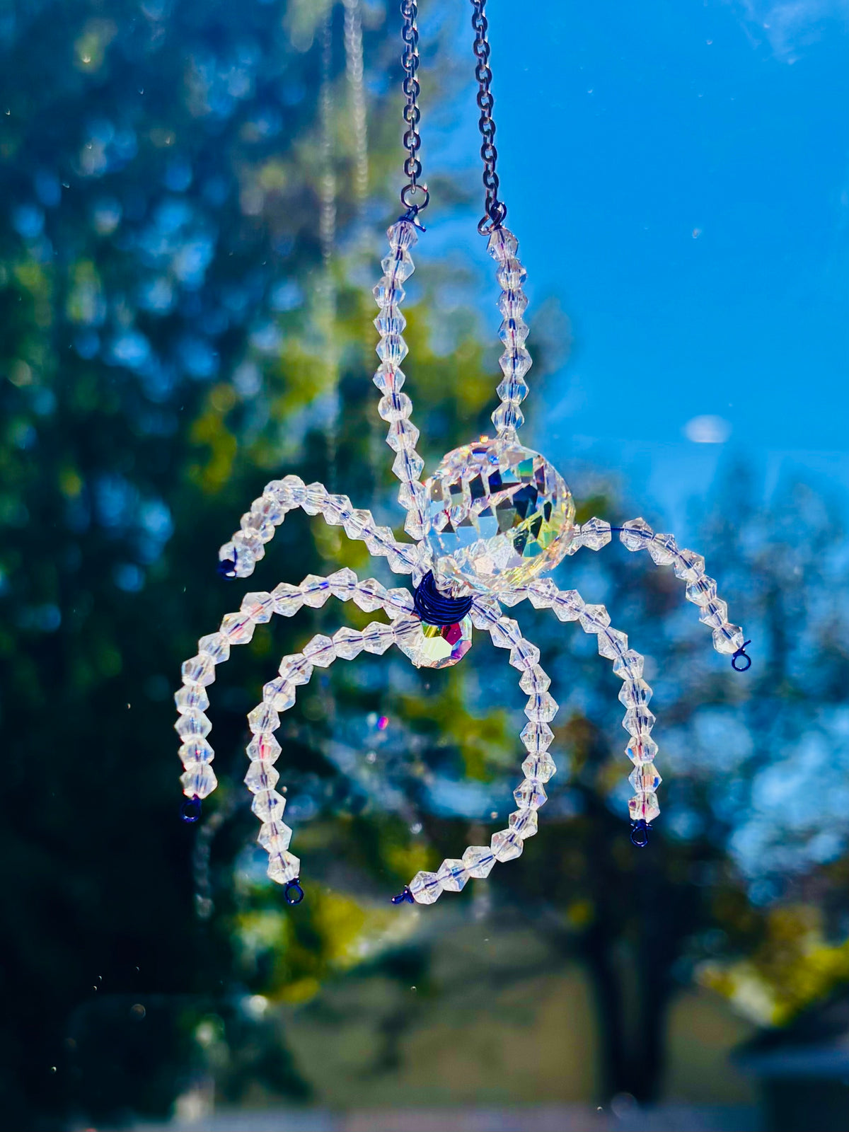 Close-up shot of a handmade Swarovski crystal spider suncatcher hanging outdoors. The faceted glass body and beaded iridescent legs shimmer brightly against a vibrant background of blue sky and sunny autumn trees.