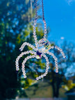 Close-up shot of a handmade Swarovski crystal spider suncatcher hanging outdoors. The faceted glass body and beaded iridescent legs shimmer brightly against a vibrant background of blue sky and sunny autumn trees.