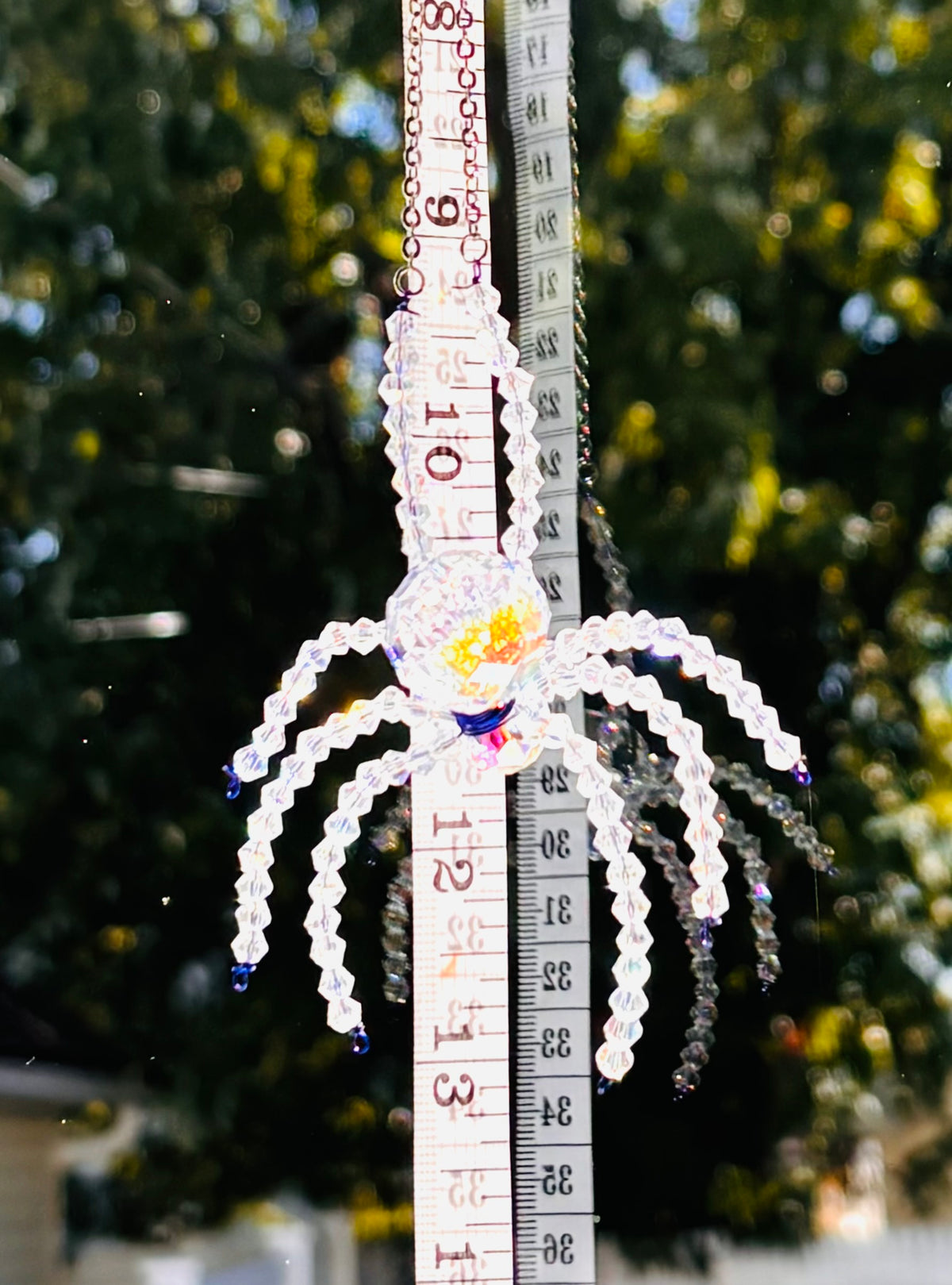 A detailed close-up shot of a handmade crystal spider suncatcher being measured with a yellow tape measure. The tape measure clearly displays the scale and size of the spider's crystal body and beaded legs against a bright outdoor backdrop.