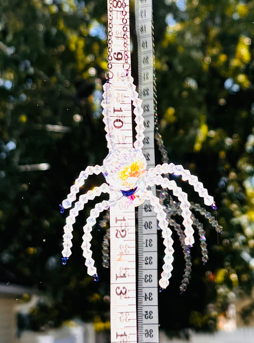 A detailed close-up shot of a handmade crystal spider suncatcher being measured with a yellow tape measure. The tape measure clearly displays the scale and size of the spider's crystal body and beaded legs against a bright outdoor backdrop.