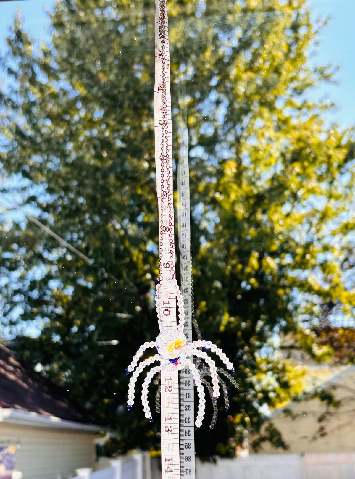 A handmade crystal spider suncatcher being measured with a yellow tape measure outdoors. The tape measure shows the precise scale and hanging length of the spider component against a background of bright autumn trees.