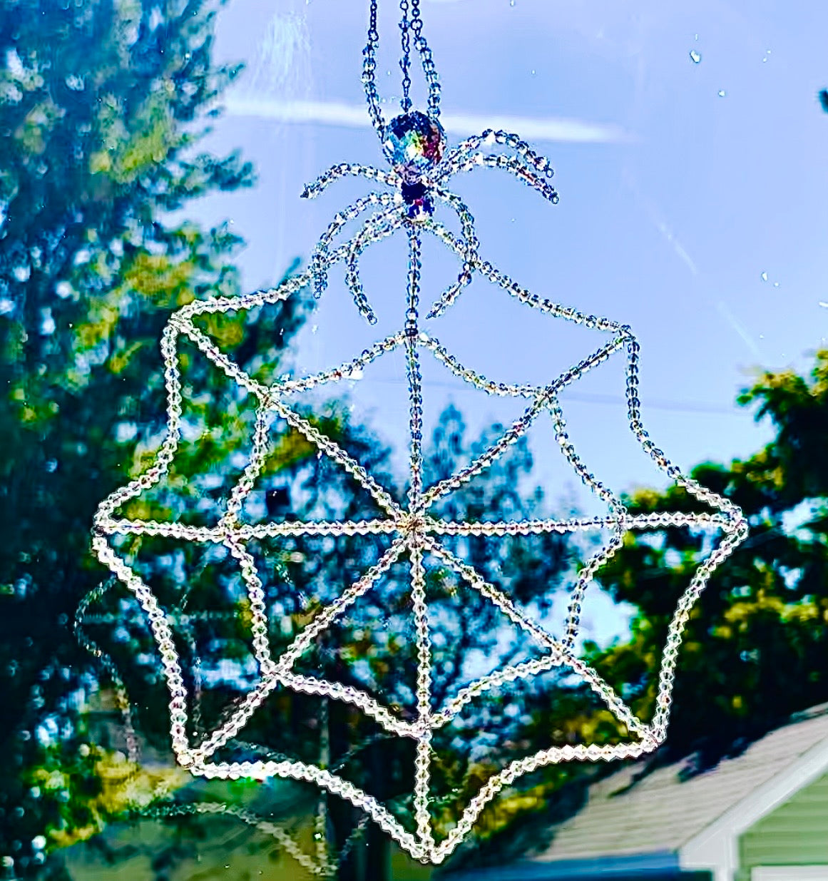 Handmade glass crystal spider and web suncatcher hanging in a sunny window against a soft-focus backdrop of green trees. The intricate beaded spiderweb is hand-threaded with 448 iridescent bicone crystals, shimmering brilliantly in the natural sunlight as a large faceted Swarovski crystal spider sits perched at the very top.