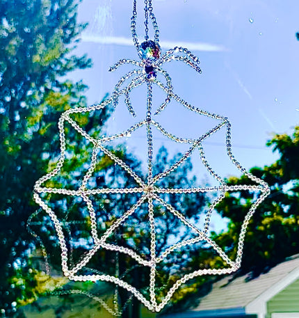 Handmade glass crystal spider and web suncatcher hanging in a sunny window against a soft-focus backdrop of green trees. The intricate beaded spiderweb is hand-threaded with 448 iridescent bicone crystals, shimmering brilliantly in the natural sunlight as a large faceted Swarovski crystal spider sits perched at the very top.