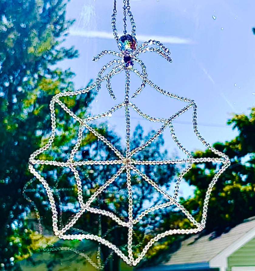 Handmade glass crystal spider and web suncatcher hanging in a sunny window against a soft-focus backdrop of green trees. The intricate beaded spiderweb is hand-threaded with 448 iridescent bicone crystals, shimmering brilliantly in the natural sunlight as a large faceted Swarovski crystal spider sits perched at the very top.