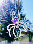 A primary product photo of a handmade Aurora Borealis ASFOUR crystal spider suncatcher hanging outdoors in crisp daylight. The large faceted crystal body gleams with intense purple and gold iridescent reflections against a soft-focus background of trees and blue sky.
