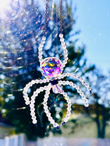 A primary product photo of a handmade Aurora Borealis ASFOUR crystal spider suncatcher hanging outdoors in crisp daylight. The large faceted crystal body gleams with intense purple and gold iridescent reflections against a soft-focus background of trees and blue sky.
