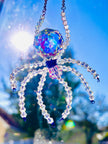 An ultra-close macro photo of an Aurora Borealis ASFOUR crystal spider suncatcher. The highly refractive faceted crystal body glints in the bright sunlight, displaying a brilliant rainbow shift of blue, purple, and magenta colors against a vivid sky.
