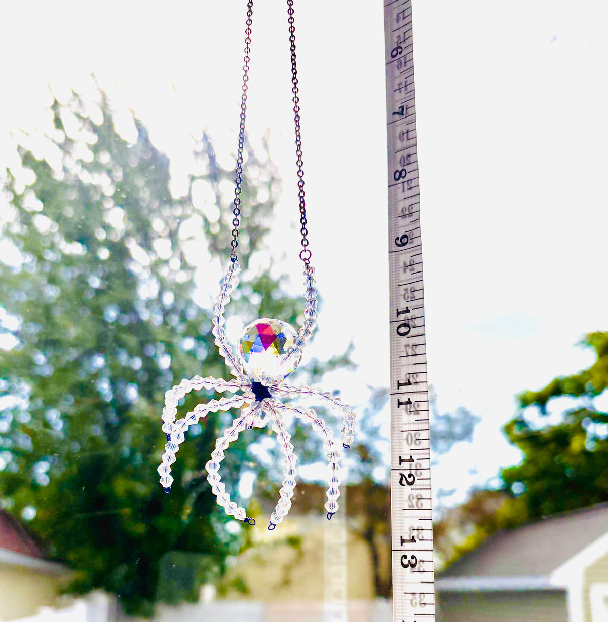 A handmade ASFOUR crystal spider suncatcher being measured vertically with a white tape measure outdoors. The tape measure shows a total hanging length of 13 inches against a bright backdrop of sky and trees.
