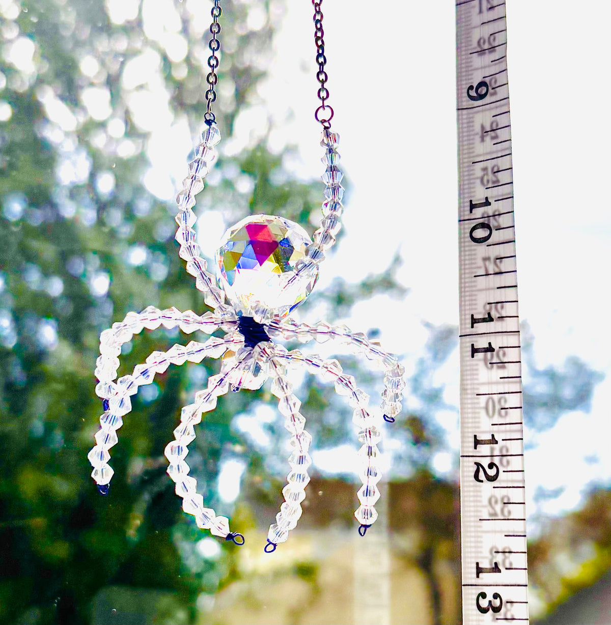 A handmade ASFOUR crystal spider suncatcher being measured vertically with a white tape measure outdoors. The tape measure shows a total spider length of about inches against a bright backdrop of sky and trees.
