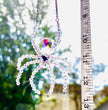 A handmade ASFOUR crystal spider suncatcher being measured vertically with a white tape measure outdoors. The tape measure shows a total spider length of about inches against a bright backdrop of sky and trees.
