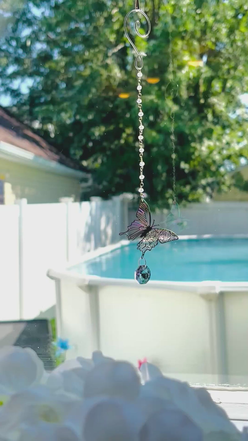 A metallic butterfly on a beaded chain spinning in a bright window with an iridescent octagonal crystal hanging from its wing. 