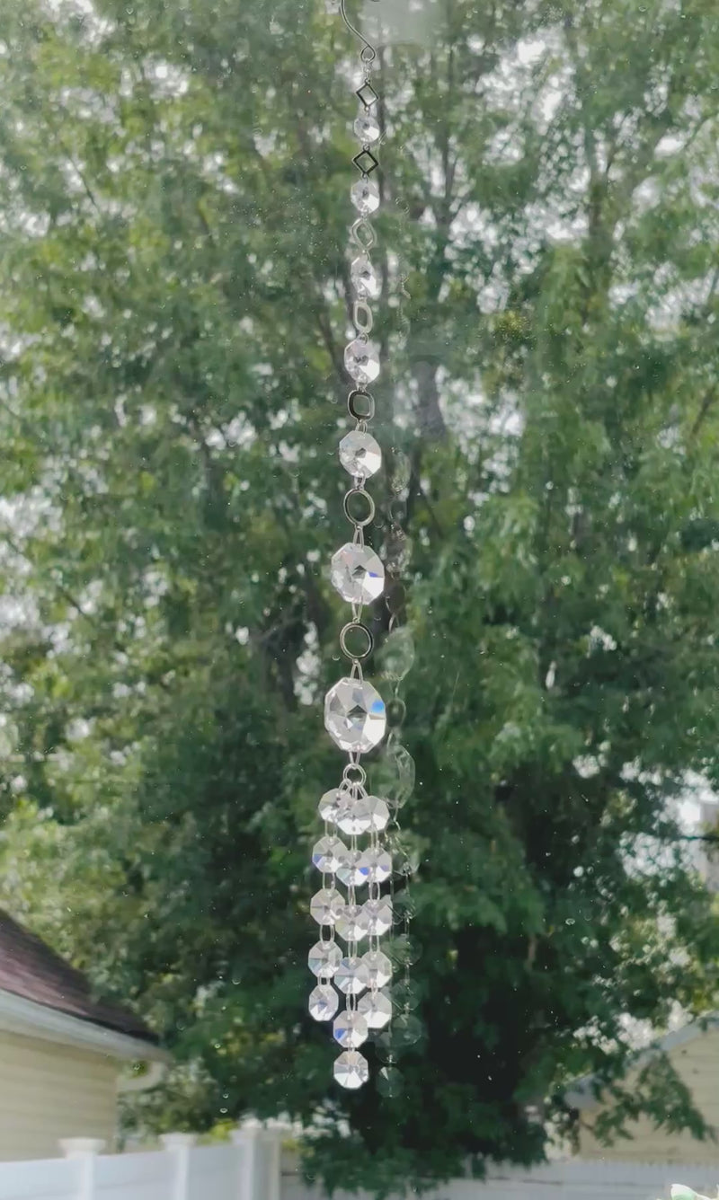 Handmade Neptune’s Octagon Prism crystal suncatcher spinning in a window against a background of lush green trees, capturing bright sunlight lighting it up through its clear faceted glass prisms.