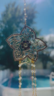 Handcrafted Winter’s Gold crystal suncatcher spinning gently in the bright sun, showcasing its intricate gold filigree flower center and shimmering crystal strands against a blue outdoor background.