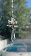 Handcrafted Hopea crystal angel suncatcher spinning gently in bright sunlight, showing a side view of its smoky metallic wings and long faceted prism body casting warm rainbow shimmers over a pool background.