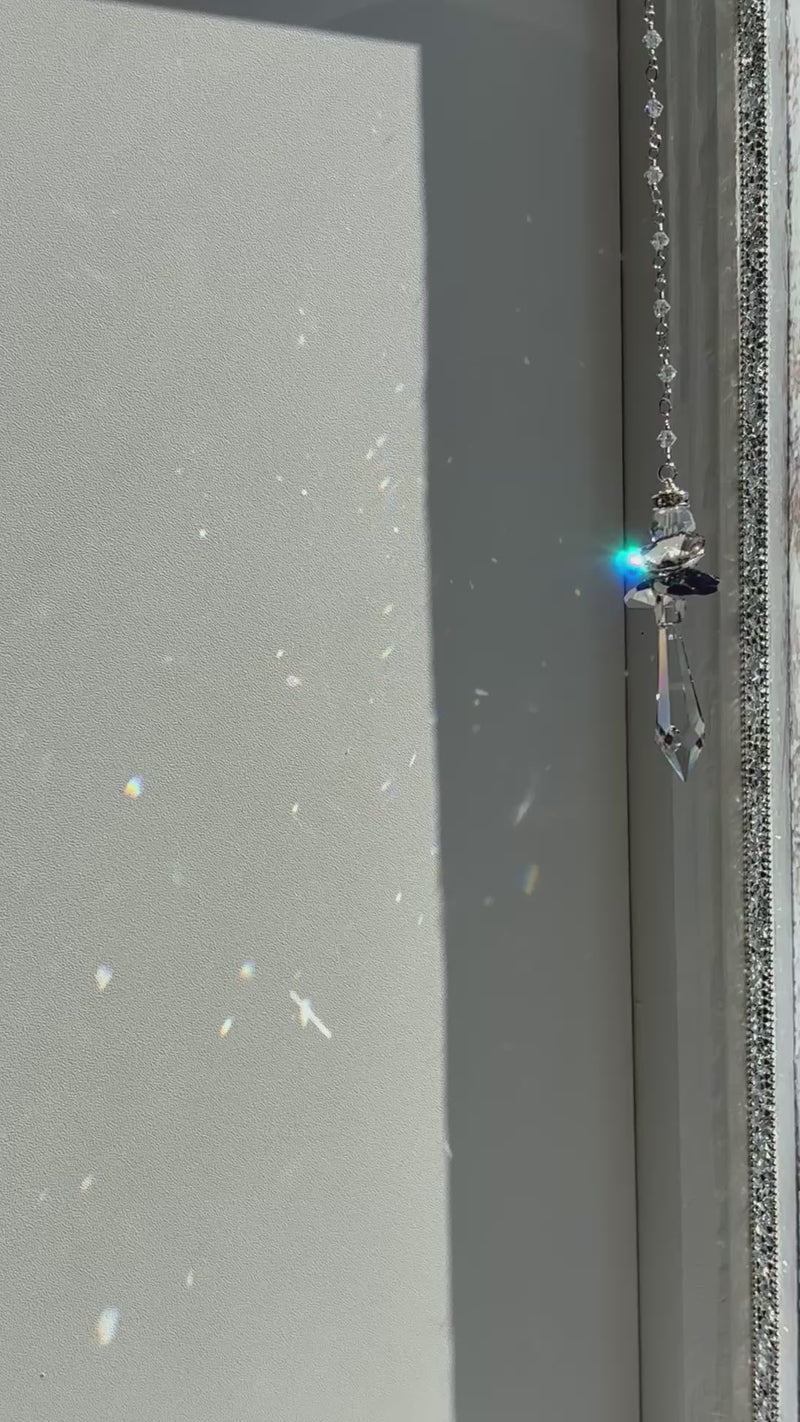 Handcrafted Hopea crystal angel suncatcher spinning gently in bright sunlight, showing a side view of its smoky metallic wings and long faceted prism body casting vibrant prisms.