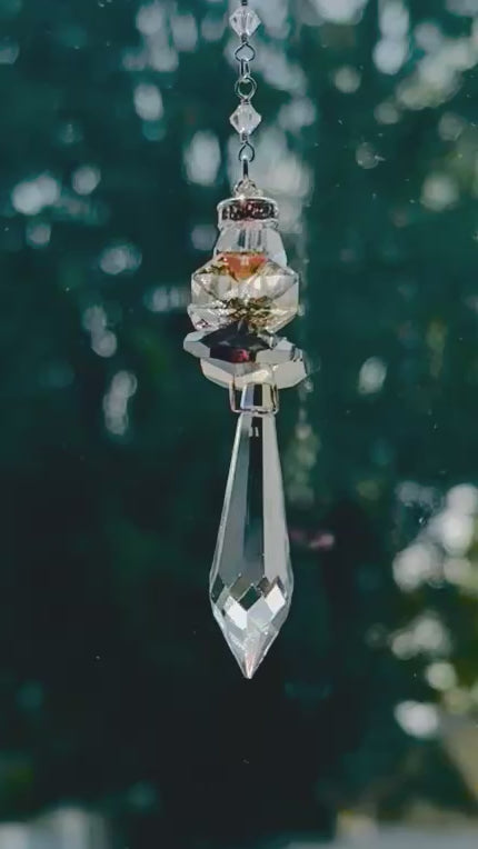 Handcrafted Hopea crystal angel suncatcher spinning gently in bright sunlight, showing a side view of its smoky metallic wings and long faceted prism body casting warm rainbow shimmers with greenery background.