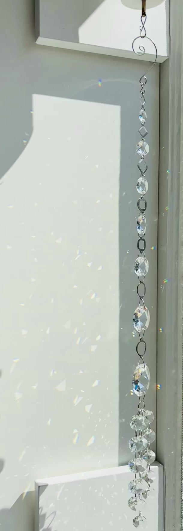 Vertical strand of Neptune's Octagon Prism crystal suncatcher hanging indoors next to a white wall covered in subtle, soft rainbow light reflections from the faceted glass prisms.