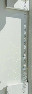Vertical strand of Neptune's Octagon Prism crystal suncatcher hanging indoors next to a white wall covered in subtle, soft rainbow light reflections from the faceted glass prisms.