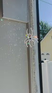 A handmade Aurora Borealis ASFOUR crystal spider suncatcher hanging in a window frame. Direct sunlight hits the faceted crystal prism and glass beads, throwing a dense pattern of bright rainbow light sparkles across the indoor wall.
