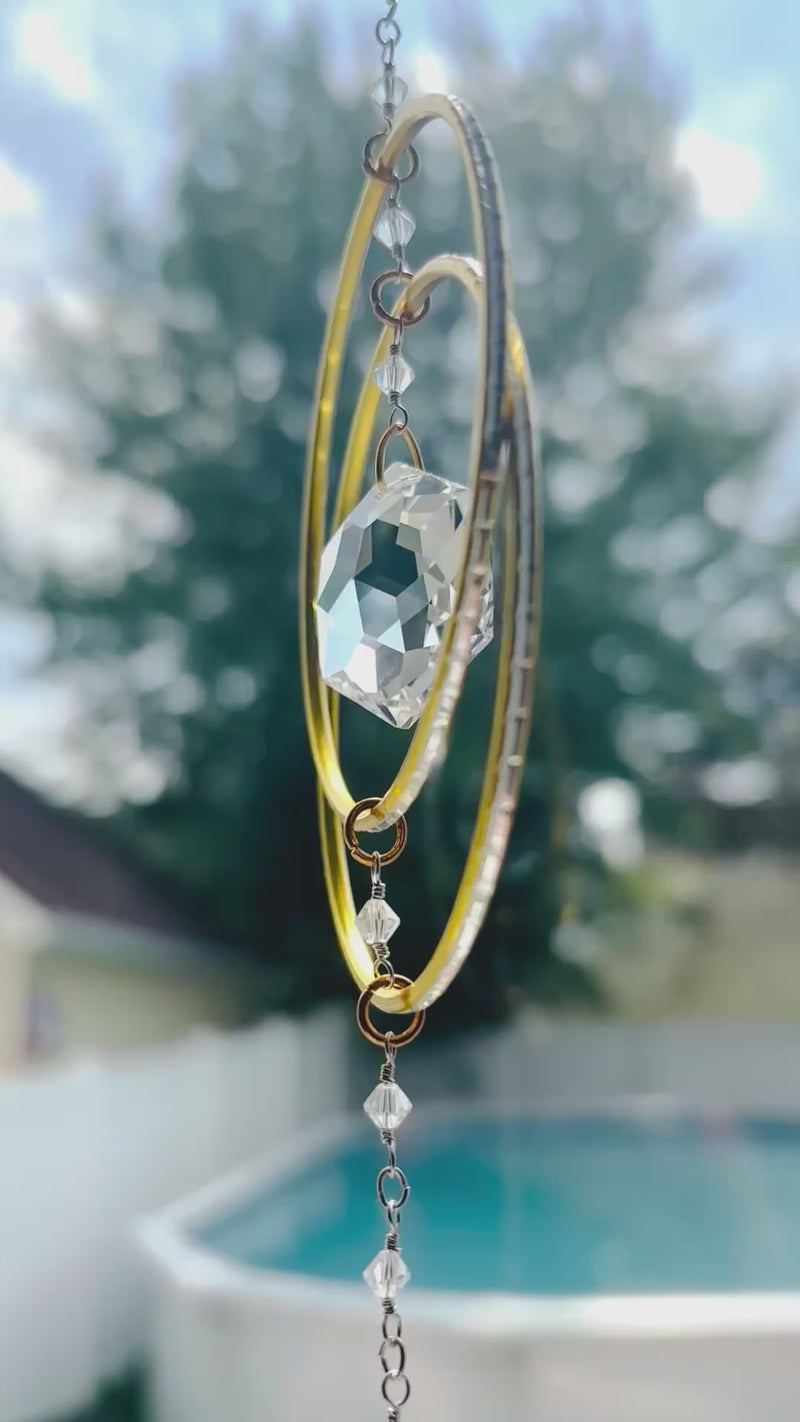 Artisan crystal suncatcher with gold hoops and a teardrop Swarovski crystal prism gently spinning in a sunny outdoor setting with a pool in the background.