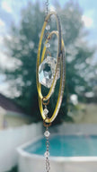 Artisan crystal suncatcher with gold hoops and a teardrop Swarovski crystal prism gently spinning in a sunny outdoor setting with a pool in the background.