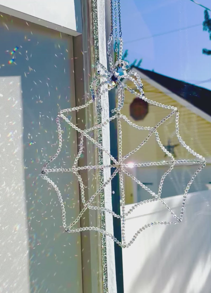 Full-length view of the Amara Collection crystal spider and web suncatcher hanging vertically next to a window frame. The natural daylight brightly illuminates the entire hand-beaded spiderweb and the detailed crystal spider, showcasing its full size and scale.