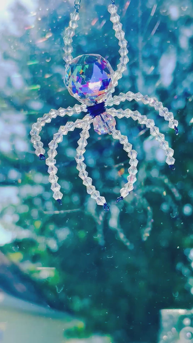 Product video of a handmade Aurora Borealis ASFOUR crystal spider suncatcher gently swaying in the natural morning light. The crystal abdomen and beaded iridescent legs shimmer with brilliant color shifts, filmed against a backdrop of a real dew-covered spiderweb in a garden.
