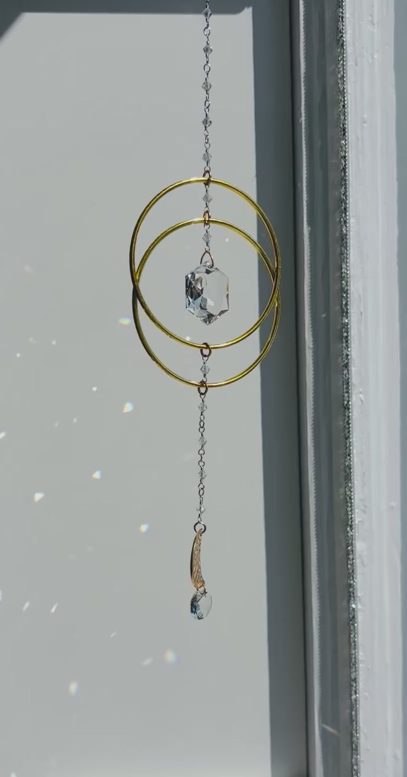Artisan crystal suncatcher with gold hoops and a teardrop Swarovski crystal prism gently spinning and creating prisms on wall as the sun hits it. 